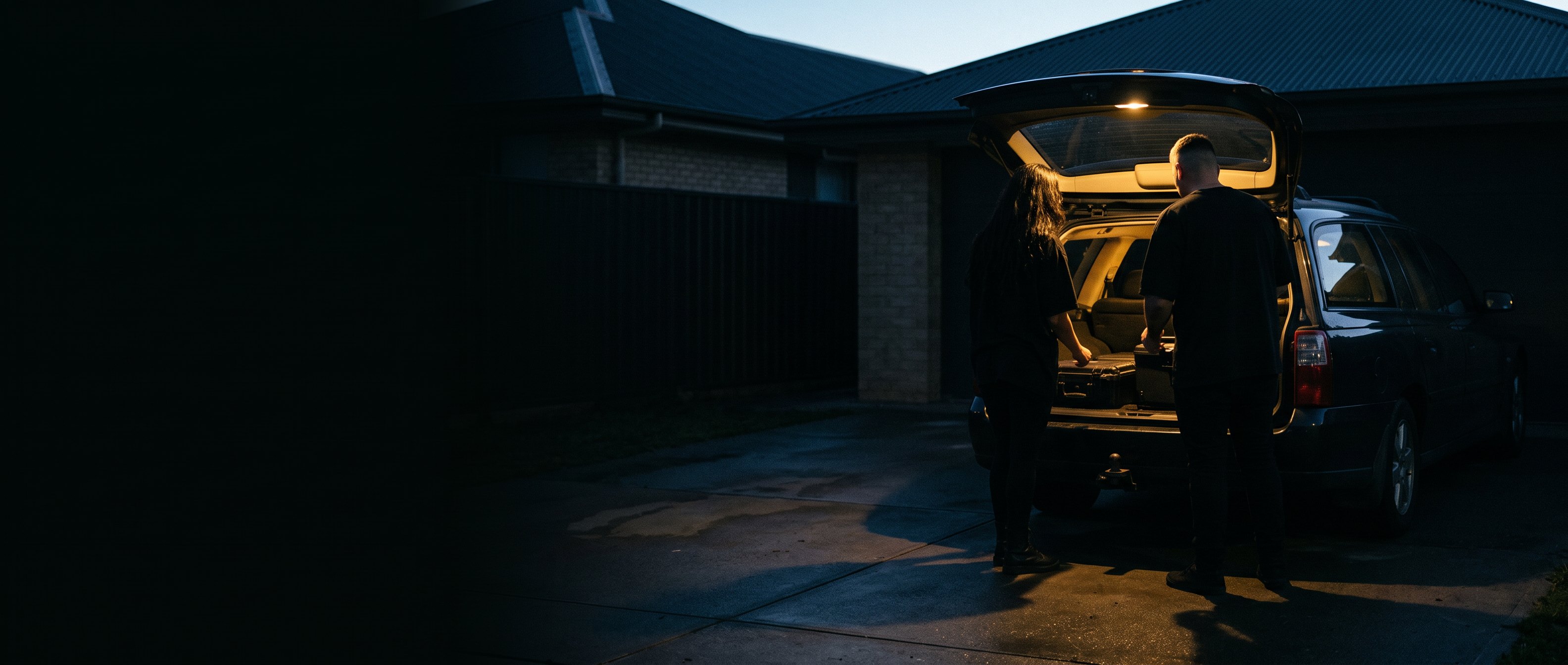 Two people loading gear into the open boot of a car in a driveway at dusk, backs to camera.