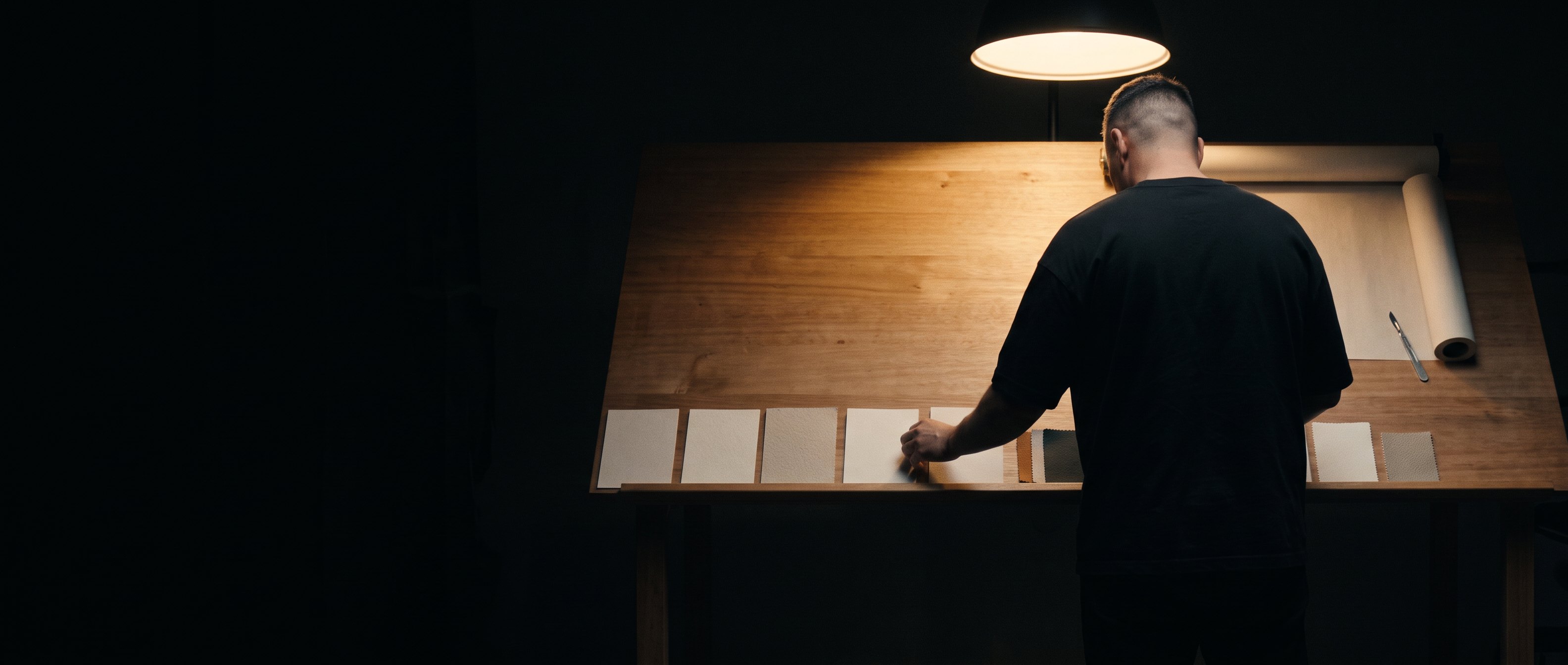 A man standing at a lit drafting table under a pendant light, back to camera, laying out printed cards.