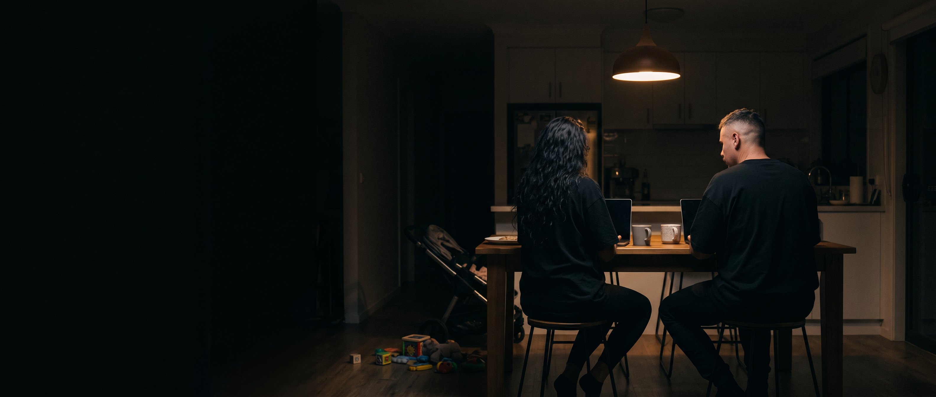 A woman and a man seated at a kitchen table under a pendant light at night, backs to camera.