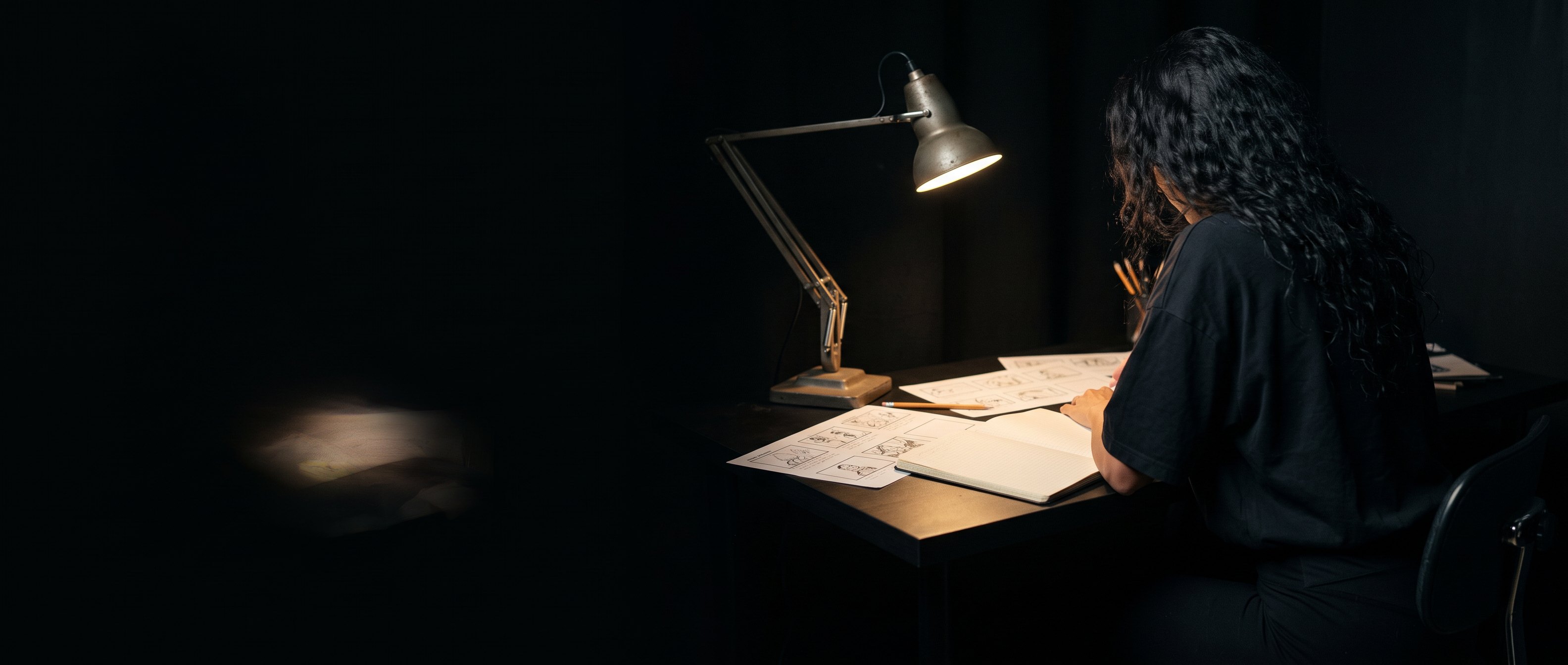 A woman with long dark hair at a desk under an angled lamp, back to camera, working over printed pages.